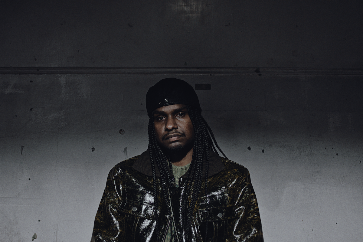 Indigenous Australian rapper Baker Boy posing in a textured jacket and cap against a dark wall in a moody promo portrait.